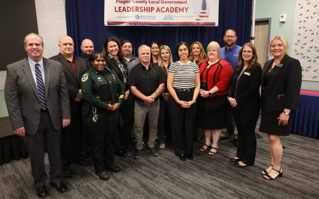 A group of people, including individuals in formal attire and one in a sheriff's uniform, pose for a photo in front of a "Flagler County Local Government Leadership Academy" banner They stand on a patterned carpeted floor in a conference room