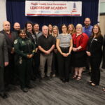 A group of people, including individuals in formal attire and one in a sheriff's uniform, pose for a photo in front of a "Flagler County Local Government Leadership Academy" banner They stand on a patterned carpeted floor in a conference room