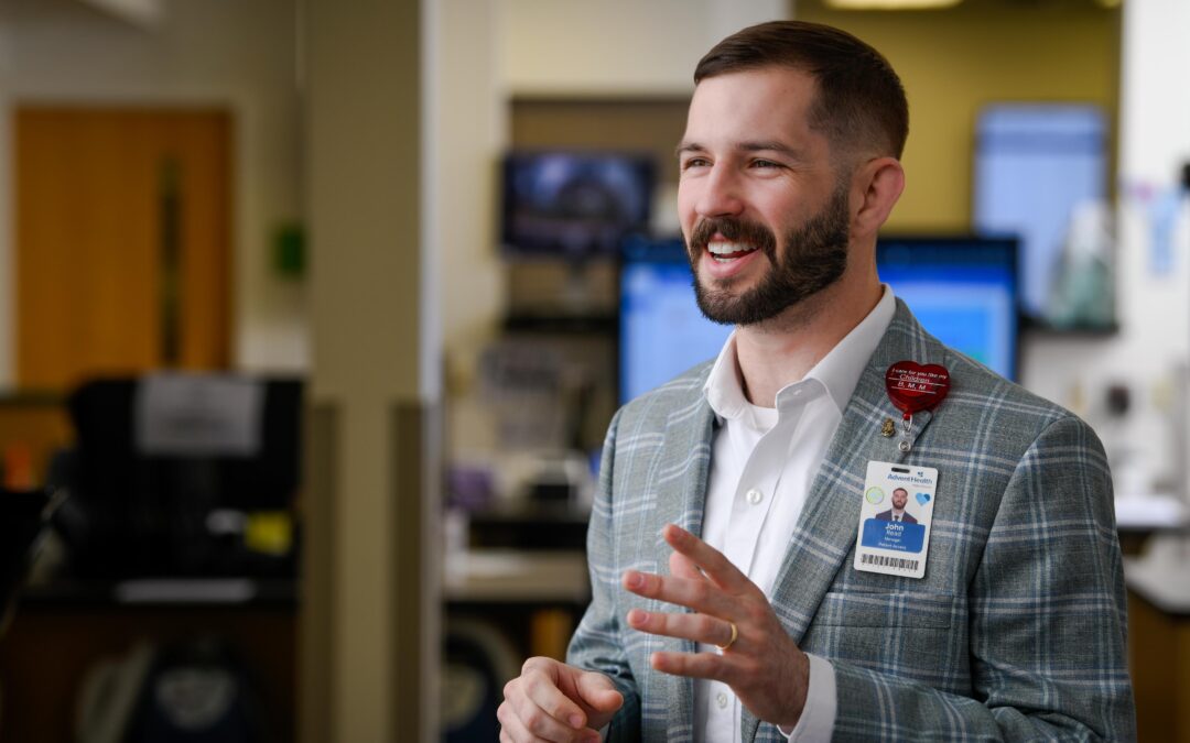 A person wearing a gray plaid suit jacket and a name badge is smiling and gesturing with hands They are standing in an office setting with computers in the background