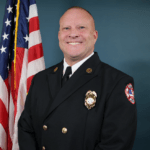 A bald man in a dark formal uniform stands smiling in front of a U S flag, against a solid blue background The uniform features gold buttons, a badge, and a fire department patch on the sleeve