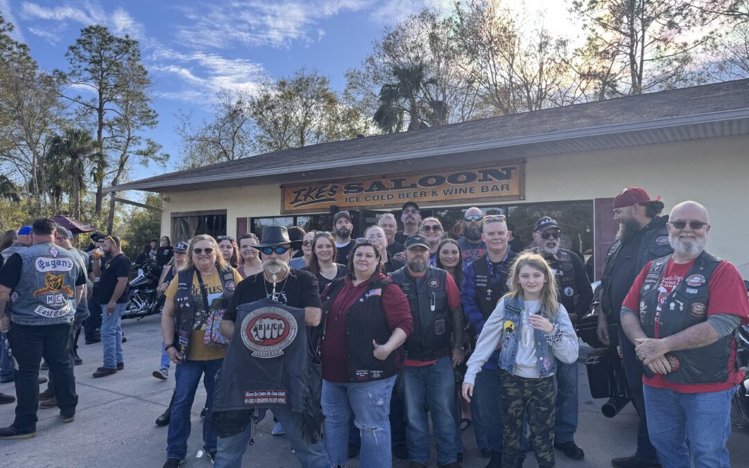 A group of people wearing biker gear stands in front of a building with a sign that reads "Ike's Saloon Ice Cold Beer & Wine Bar " They appear to be posing for a photo on a sunny day, with trees in the background