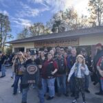 A group of people wearing biker gear stands in front of a building with a sign that reads "Ike's Saloon Ice Cold Beer & Wine Bar " They appear to be posing for a photo on a sunny day, with trees in the background