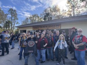 A group of people wearing biker gear stands in front of a building with a sign that reads "Ike's Saloon Ice Cold Beer & Wine Bar " They appear to be posing for a photo on a sunny day, with trees in the background