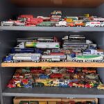 A collection of toy model vehicles is displayed on four shelves The assortment includes tankers, trucks, cars, and emergency vehicles in various colors and designs, with brand names visible on some The shelves are made of dark wood