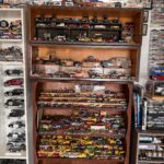 A collection of toy cars displayed on shelves The shelves are filled with various models, including vintage and construction vehicles The background showcases framed images and additional toy cars