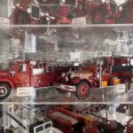 A display case containing two red vintage fire truck models The front truck is labeled " GMC" and the rear truck is labeled " Stoughton " Other model fire trucks are visible in the background