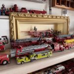 A collection of various fire truck models displayed on shelves The models are red and include ladder trucks, with some smaller yellow trucks A gold framed picture is partly visible in the background Toy figurines are arranged around the trucks