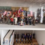 A collection of miniature military figurines displayed in a cabinet The figures are dressed in various historical uniforms, holding flags, and arranged on multiple shelves A book is partially visible on the lower shelf