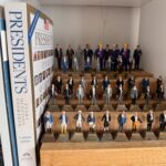 A collection of small U S president figurines arranged in rows on a shelf, alongside two books titled "Presidents" and "Presidents: Every Question Answered " Another shelf with more figurines is partially visible