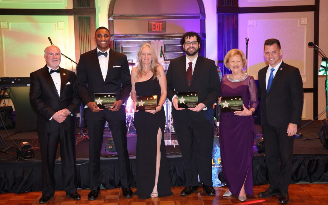 Six people dressed formally stand on a wooden floor, holding plaques The backdrop includes musical instruments and colorful lighting