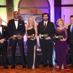 Six people dressed formally stand on a wooden floor, holding plaques The backdrop includes musical instruments and colorful lighting