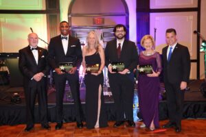 Six people dressed formally stand on a wooden floor, holding plaques The backdrop includes musical instruments and colorful lighting