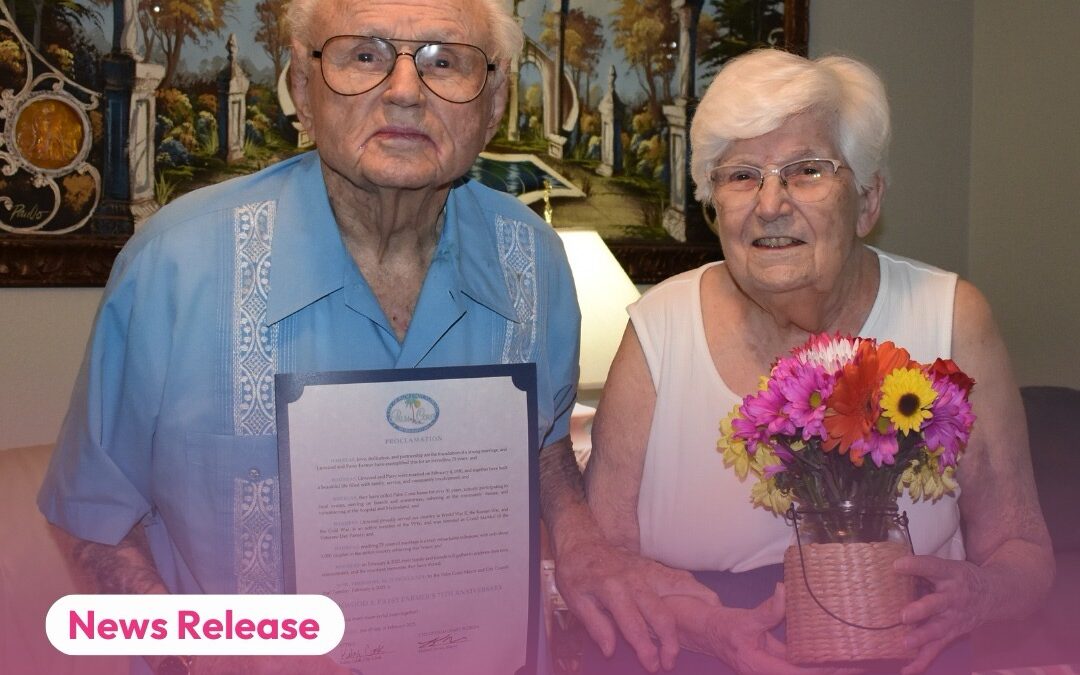 An elderly couple, both wearing glasses, holding a framed city proclamation The man is in a light blue shirt, and the woman in a white dress, holding a bouquet of flowers They're in a room with a landscape painting behind them