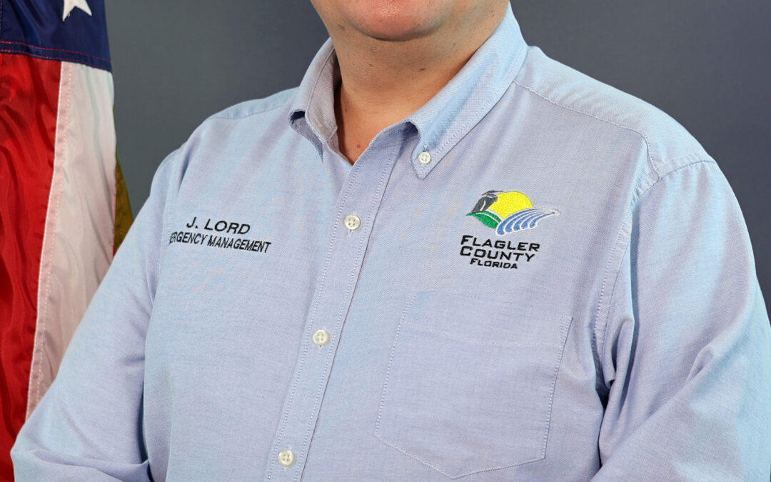 A man with short dark hair stands in front of a dark background, wearing a light blue shirt with "Emergency Management" and a logo on it An American flag is visible to his left