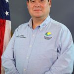 A man with short dark hair stands in front of a dark background, wearing a light blue shirt with "Emergency Management" and a logo on it An American flag is visible to his left