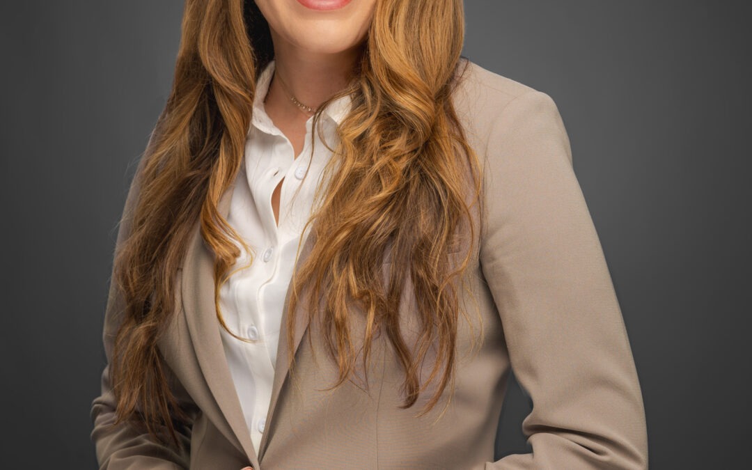 Woman with long brown hair wearing a beige suit and white blouse smiles, posing against a dark grey background