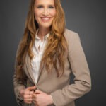 Woman with long brown hair wearing a beige suit and white blouse smiles, posing against a dark grey background