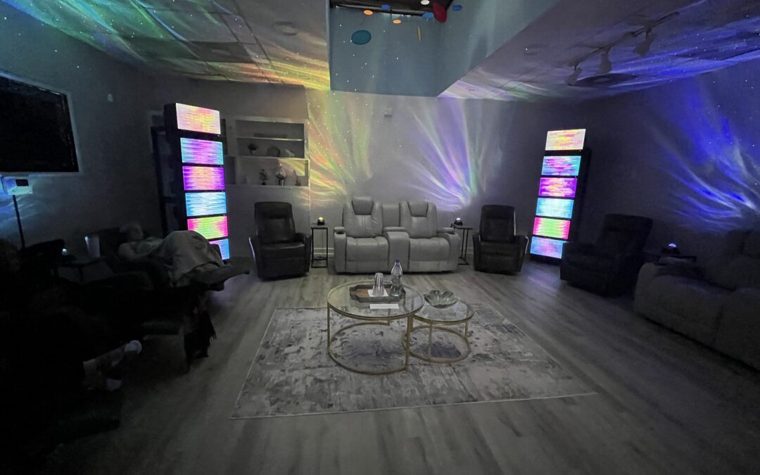 A dimly lit room with multiple recliner chairs arranged in a semicircle Colorful, abstract light patterns are projected on the walls A rug and two coffee tables are in the center, creating a cozy, ambient atmosphere