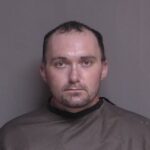 A man with short hair and a trimmed beard looks directly at the camera He is wearing a dark drape, typical of mugshots, against a plain gray background