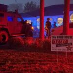 A nighttime scene with police activity outside a house A sign in the yard reads "This drug house closed for business by Sheriff Rick Stacy " A police vehicle and several officers are visible under red and blue lights