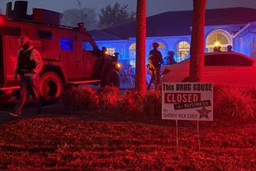 A nighttime scene with police activity outside a house A sign in the yard reads "This drug house closed for business by Sheriff Rick Stacy " A police vehicle and several officers are visible under red and blue lights