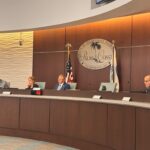 Four people sit behind a long, curved desk at a Palm Coast city council meeting in Florida An American flag and a city emblem are visible on the wall behind them Tables with microphones are in front of each person