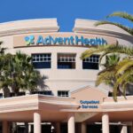 A modern beige building with palm trees in front and "AdventHealth" signage on top The structure has rounded architecture with multiple windows and a sign for "Outpatient Services" at the entrance The sky is clear and blue