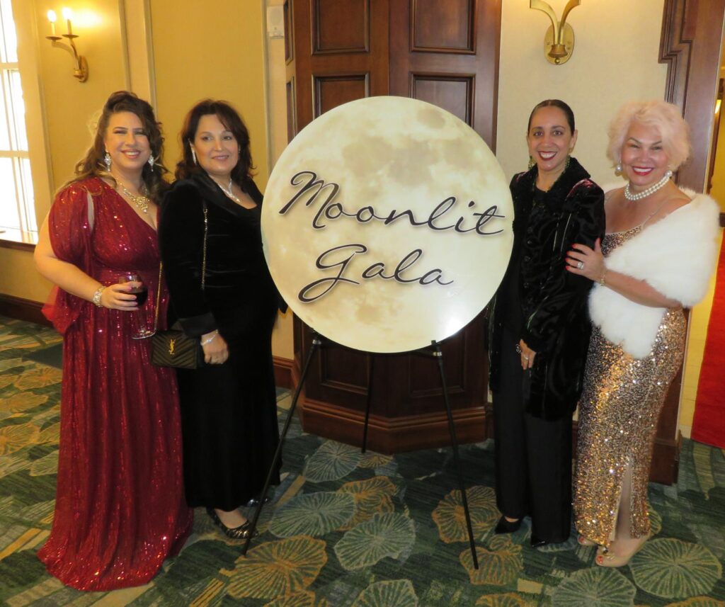 Four women in elegant dresses stand smiling by a "Moonlit Gala" sign The setting is a well lit indoor venue with carpeted flooring and a wooden door in the background