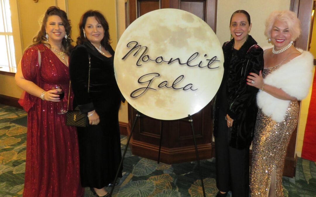 Four women in elegant dresses stand smiling by a "Moonlit Gala" sign The setting is a well lit indoor venue with carpeted flooring and a wooden door in the background