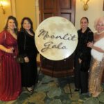 Four women in elegant dresses stand smiling by a "Moonlit Gala" sign The setting is a well lit indoor venue with carpeted flooring and a wooden door in the background