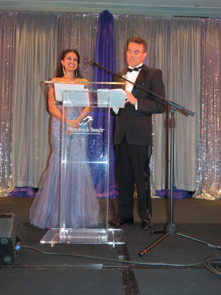 A woman and man stand at a clear podium on stage, dressed in formal evening wear The woman is in a purple gown, and the man is in a black tuxedo Behind them are shiny silver curtains, and the podium has "Hammock Beach" written on it