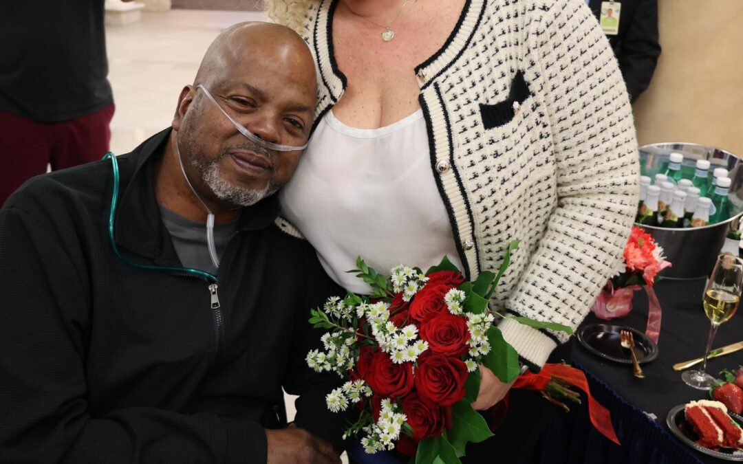 A woman with curly hair holds a bouquet of red roses and white flowers, smiling beside a seated man with an oxygen tube He leans on her, smiling A table with drinks and strawberries is nearby, and people are in the background