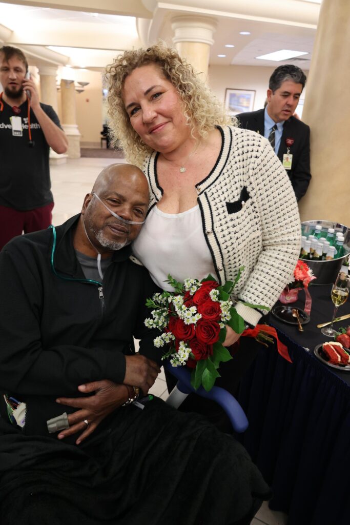 A woman with curly hair holds a bouquet of red roses and white flowers, smiling beside a seated man with an oxygen tube He leans on her, smiling A table with drinks and strawberries is nearby, and people are in the background