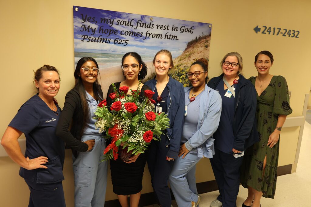 A group of seven women, some in medical scrubs, stand smiling in a hallway One woman holds a bouquet of red roses A wall poster behind them displays a Bible verse from Psalms : about finding rest and hope in God