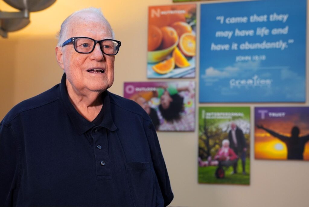 An older man wearing glasses and a dark polo shirt stands in front of a wall with vibrant posters featuring fruit, people, and inspirational quotes