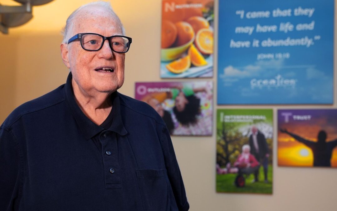 An older man wearing glasses and a dark polo shirt stands in front of a wall with vibrant posters featuring fruit, people, and inspirational quotes