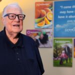 An older man wearing glasses and a dark polo shirt stands in front of a wall with vibrant posters featuring fruit, people, and inspirational quotes