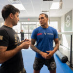 Two men are standing in a martial arts training facility, having a conversation One is wearing a black shirt and the other a blue UFC shirt The room has a padded floor and a chain link fence; other people are visible in the background