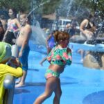 Children play in a splash pad with water sprays A girl in a floral swimsuit walks through the water, while another child in a yellow outfit and hat looks on Other children are in the background enjoying the sunny day