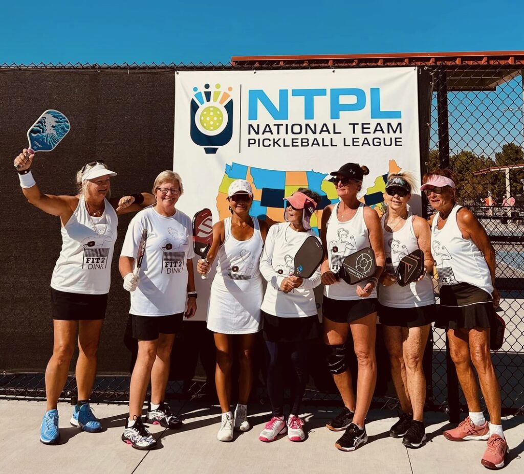 A group of seven women in athletic wear stand in front of an "NTPL National Team Pickleball League" banner They are holding paddles and smiling, with a sunny outdoor court setting in the background