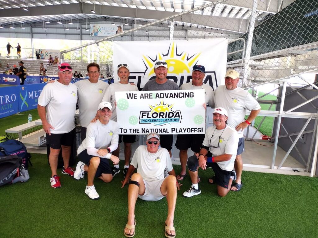 A group of nine people in white shirts and shorts are posing indoors on a sports field They are holding a banner that reads, "We are advancing Florida Pickleball League to the National Playoffs!