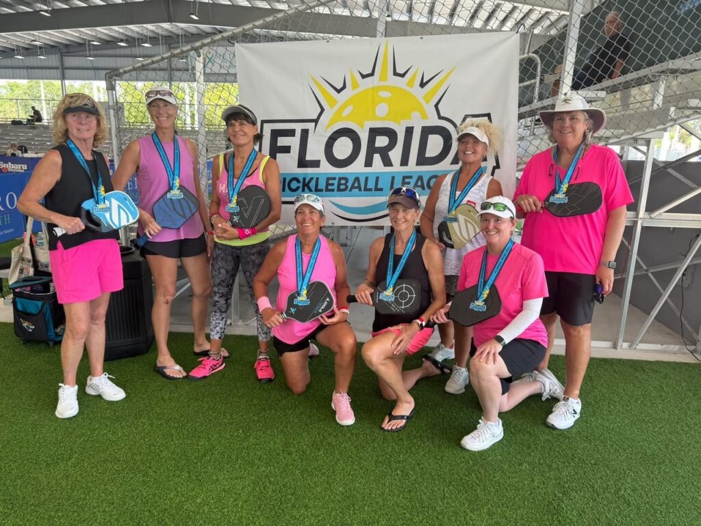 A group of nine women smiling and posing indoors with pickleball paddles They are wearing medals around their necks A banner behind them reads "Florida Pickleball League " Most are dressed in pink outfits and hats