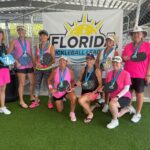 A group of nine women smiling and posing indoors with pickleball paddles They are wearing medals around their necks A banner behind them reads "Florida Pickleball League " Most are dressed in pink outfits and hats