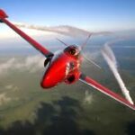 A sleek, red jet aircraft performing an acrobatic maneuver, emitting white smoke trails from its wings against a backdrop of green landscape and blue sky The plane is tilted with one wing higher, showcasing its agility