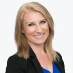 A woman with shoulder length blonde hair is smiling, wearing a black blazer over a blue top The background is plain white