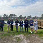 A group of eight people standing outdoors, all holding a long blue ribbon ready to be cut They are in a grassy area near a wooden deck with trees and a pond in the background The sky is cloudy