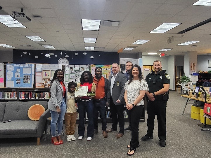 A group of people stands in a library A woman in the center holds a certificate, smiling A police officer and several others are beside her Bookshelves and posters are visible in the background