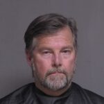 A middle aged man with gray hair and a beard is facing the camera with a neutral expression, set against a plain gray background He is wearing a dark top