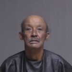 Elderly man with a mustache, wearing a dark cape like garment, stares directly at the camera against a plain gray background