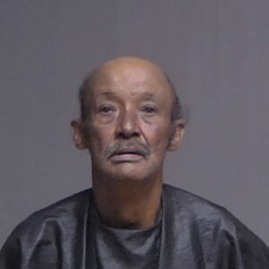 Elderly man with a mustache, wearing a dark cape like garment, stares directly at the camera against a plain gray background